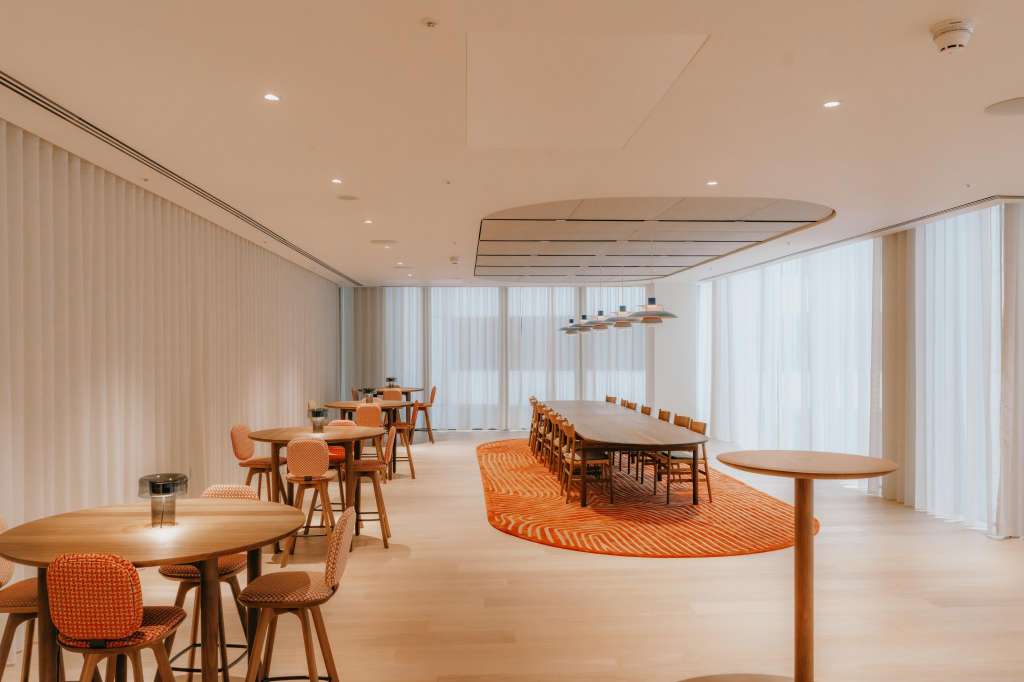 Atrium space with multiple wooden tables, orange chairs, striped rugs, and floor-to-ceiling windows with sheer curtains.