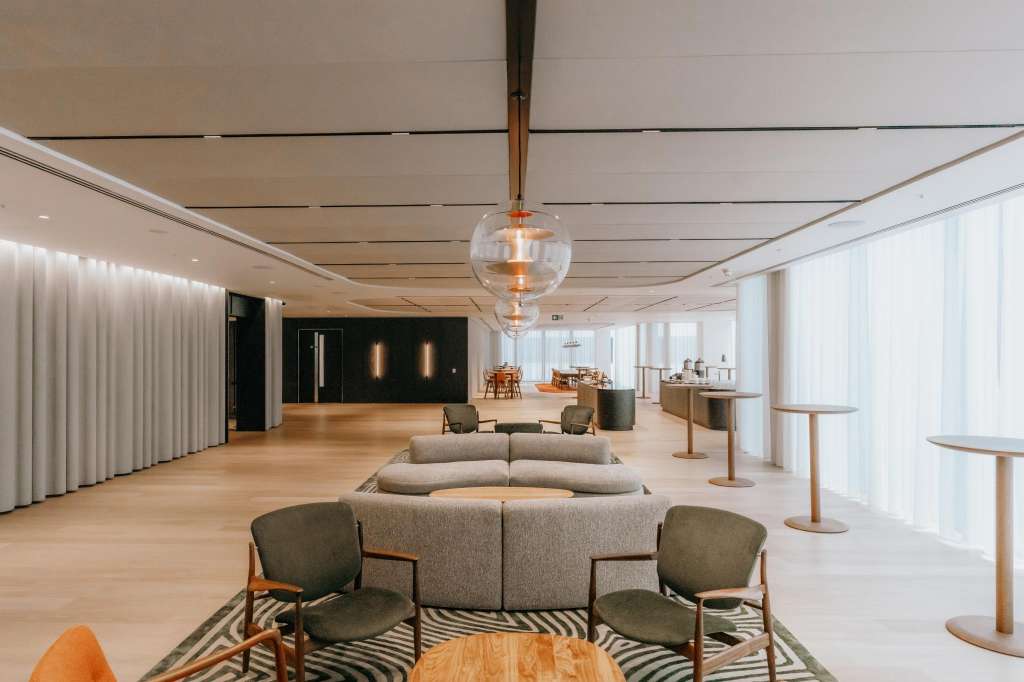 Large open atrium with modern seating areas, pendant lighting, wooden flooring, and floor-to-ceiling windows.