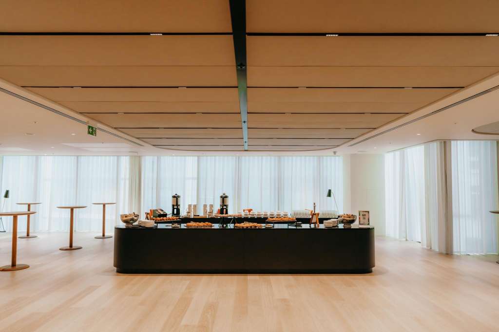 Atrium with black catering counter displaying food and beverages, high ceilings, white curtains, wooden flooring, and standing tables.