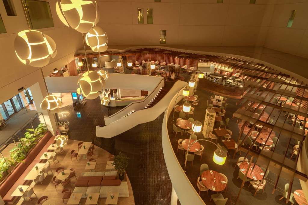 Multi-level atrium with modern pendant lighting, wooden tables, chairs, and retail displays across tiered floors.