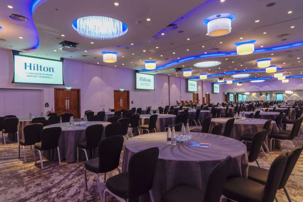 Ballroom set up with round tables, black chairs, purple linens, and modern blue-accented ceiling lighting.