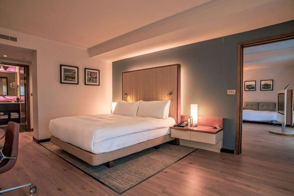 Modern hotel bedroom with king bed, wood headboard, ambient lighting, and ensuite bathroom visible through open door.