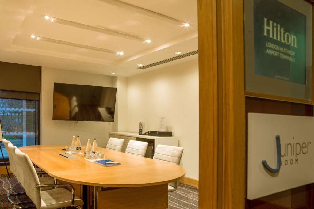 Meeting room with oval wooden table, upholstered chairs, recessed ceiling lights, and wall-mounted screens.