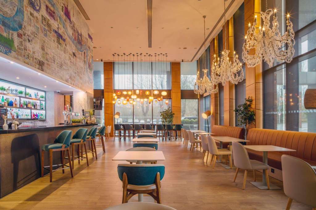 Modern lounge with bar, crystal chandeliers, wooden seating, and floor-to-ceiling windows overlooking grounds.