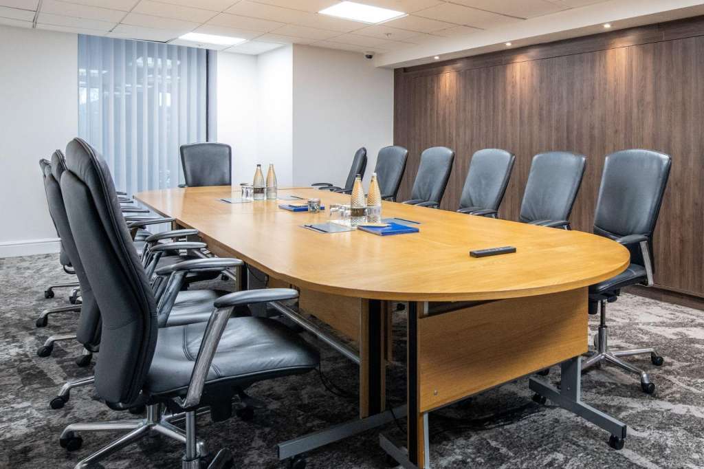 Conference room with oval wooden table, gray upholstered chairs, water bottles, notepads, and wood-paneled walls.