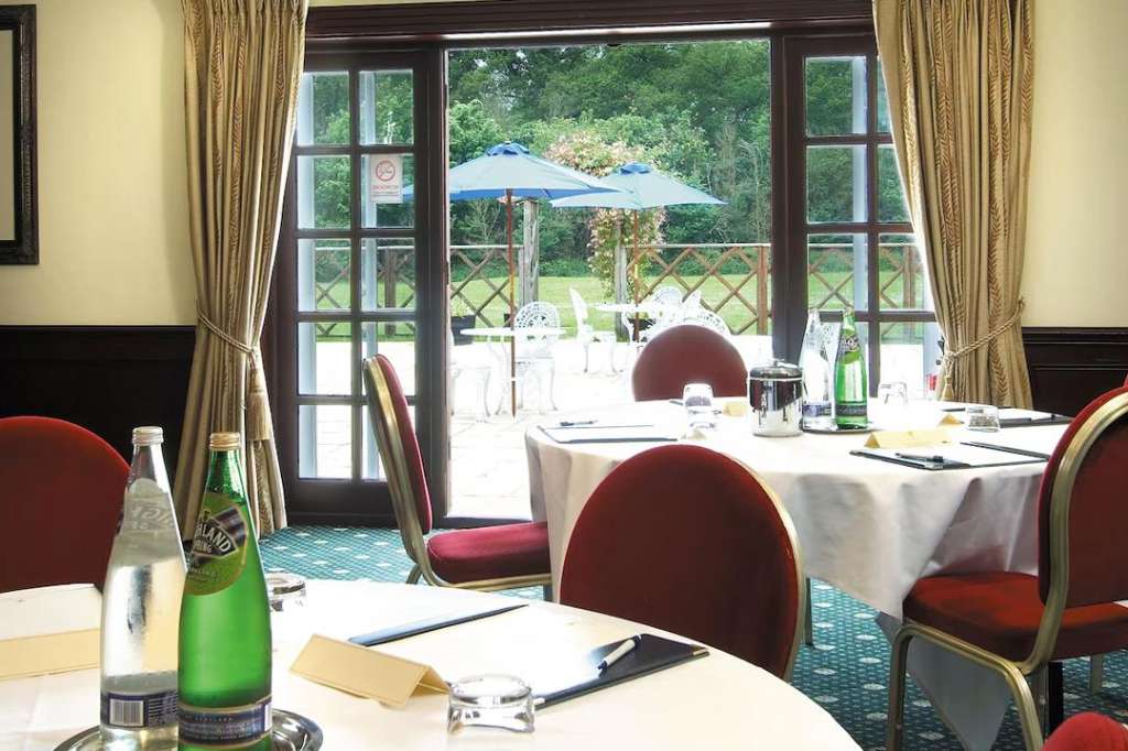 Meeting room with round tables, red chairs, notepads, and water bottles. French doors open to summer garden with blue umbrella.