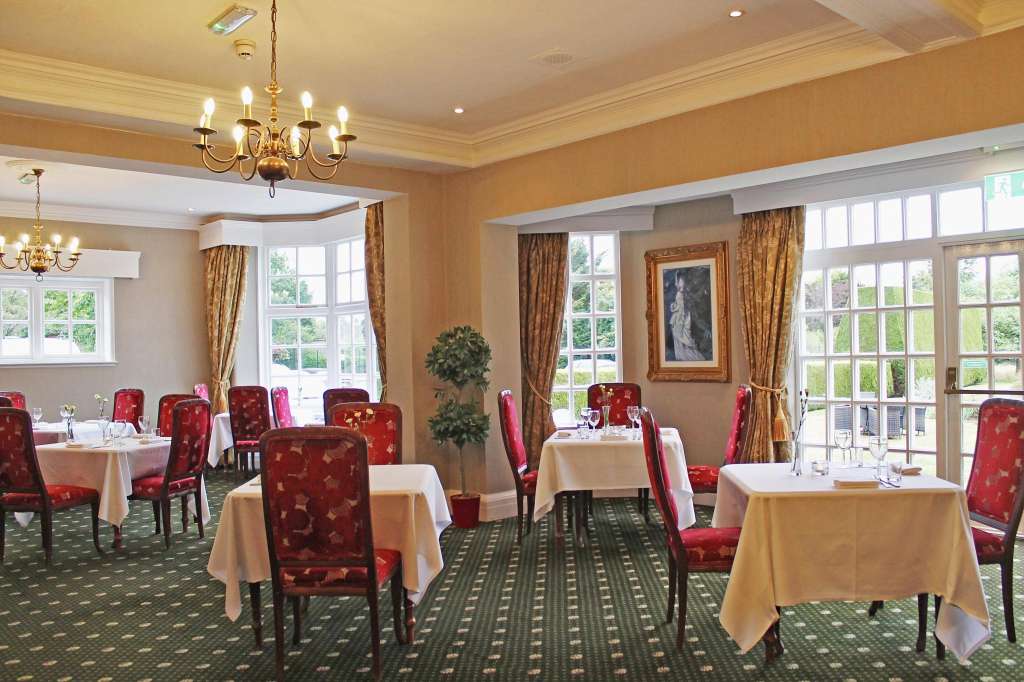 Dining room with round tables dressed in cream linens, red upholstered chairs, gold chandeliers, and large windows overlooking gardens.