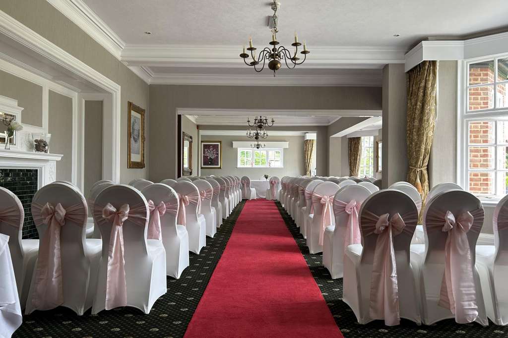 Elegant ballroom set for a formal event with white chair covers, pink sashes, red carpet aisle, and chandeliers.