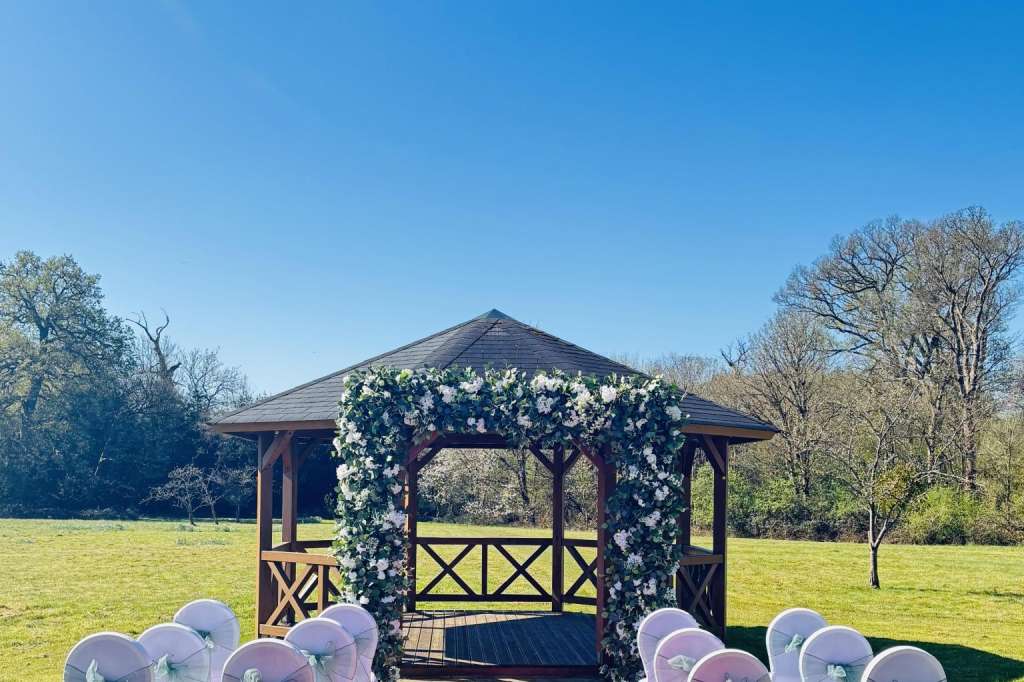 Outdoor garden ceremony setup with white chair covers, red aisle runner, and floral-draped wooden gazebo.