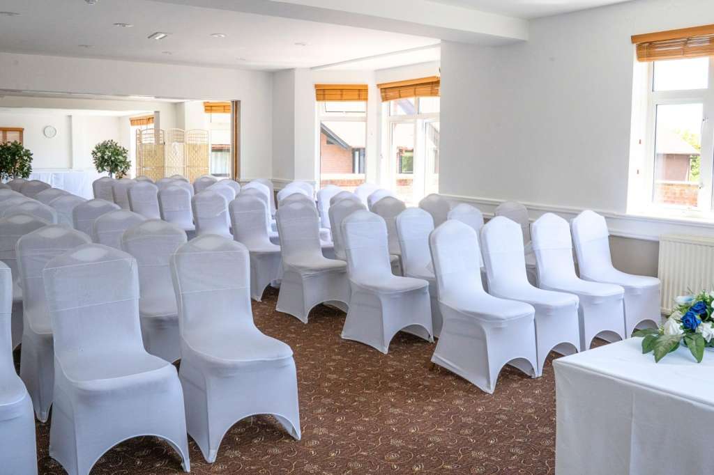Bright room with rows of white chair covers arranged theater-style, patterned carpet, large windows with orange blinds, and floral arrangement.