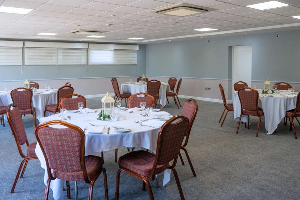 Banquet room with round tables dressed in white linens, burgundy chairs, place settings, and water glasses.