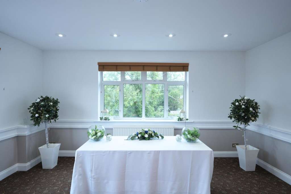 Elegant room with round table draped in white linen, flanked by topiary arrangements and large windows overlooking greenery.