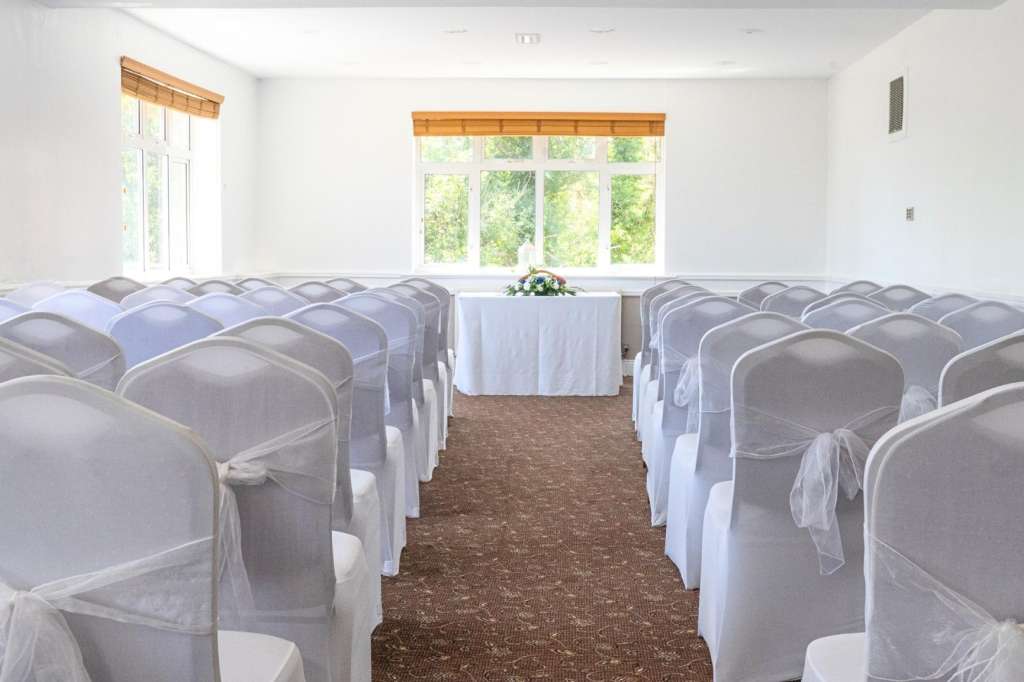 Meeting room with rows of white-draped chairs facing a small table with floral arrangement and large windows.