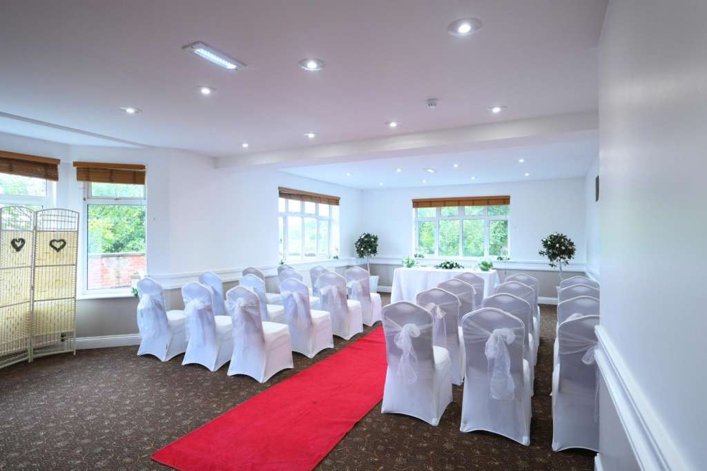 Bright room with rows of white chair covers, red aisle runner, and large windows overlooking greenery.