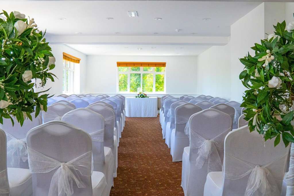 Bright room with rows of white-draped chairs facing a central aisle, flanked by potted white flowers and greenery.