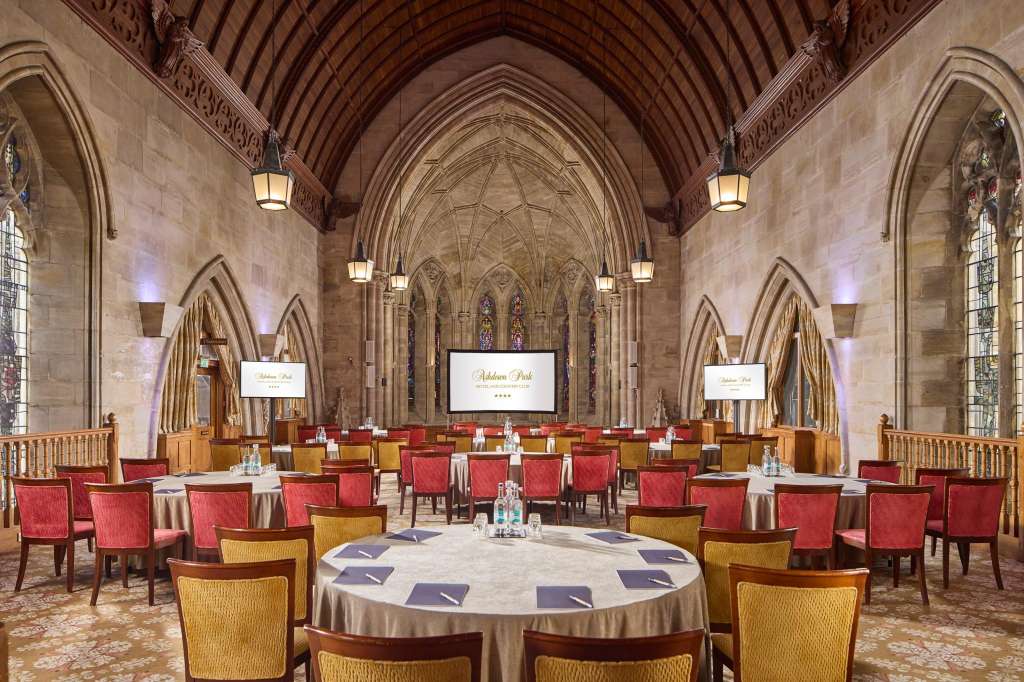 Grand hall with vaulted Gothic ceiling, round tables with red chairs, and stained glass windows.