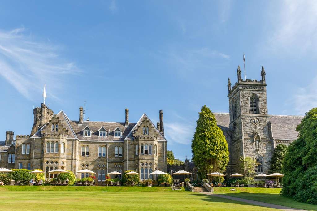Exterior view of stone manor house with manicured lawn, umbrellas, and guests on grounds.