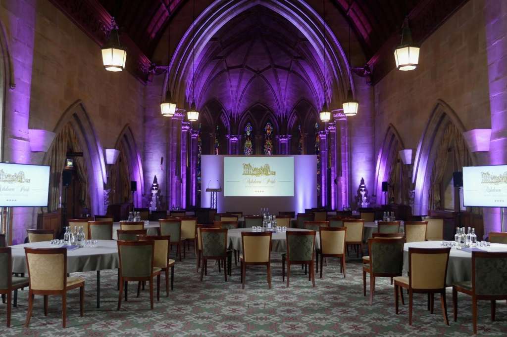 Grand hall with vaulted Gothic ceiling, purple uplighting, round tables with chairs, projection screens, and pendant lighting.