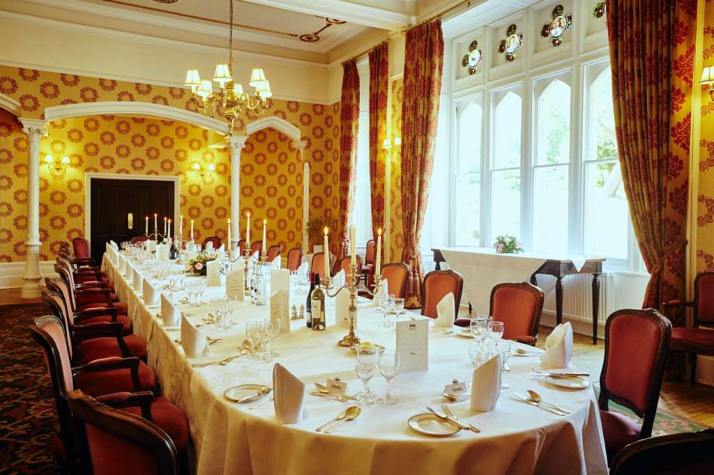 Formal dining room with long table dressed in cream linens, gold place settings, candelabras, and burgundy upholstered chairs.