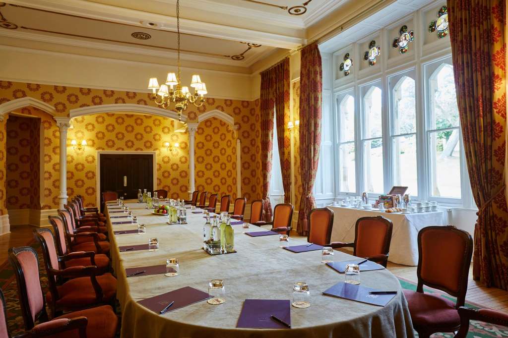Elegant meeting room with long banquet table, burgundy chairs, gold patterned wallpaper, and large windows.
