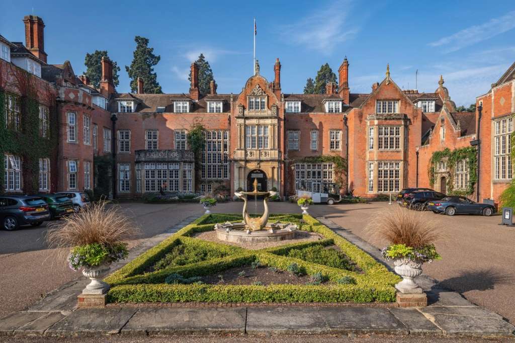 Exterior courtyard of red brick manor house with manicured gardens, fountain, and parked vehicles.