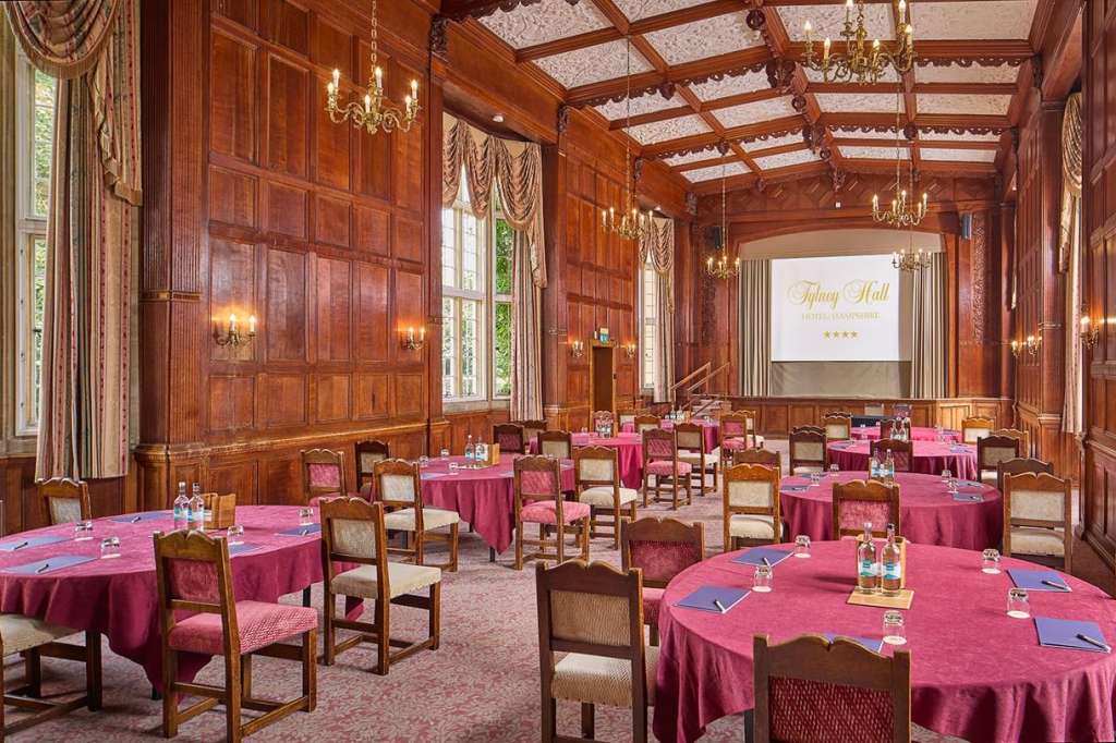Elegant wood-paneled room with round tables dressed in pink linens, gold chandeliers, and projection screen.