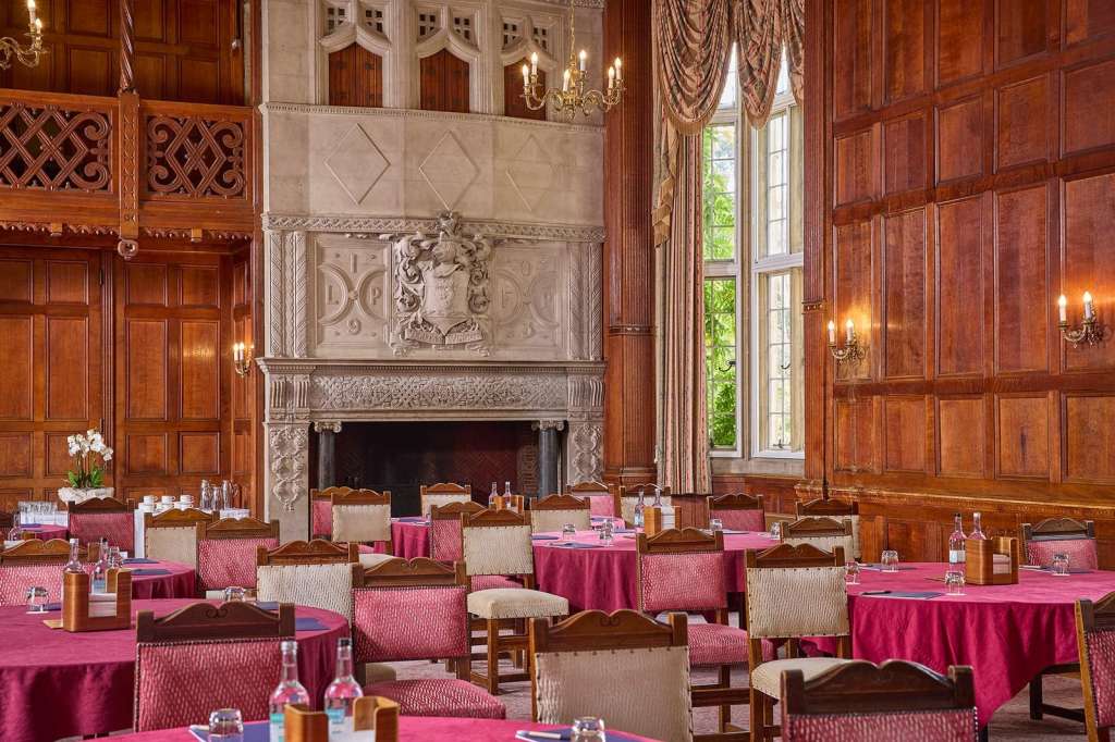 Formal dining room with ornate fireplace, wood paneling, round tables dressed in pink linens, and tall windows.