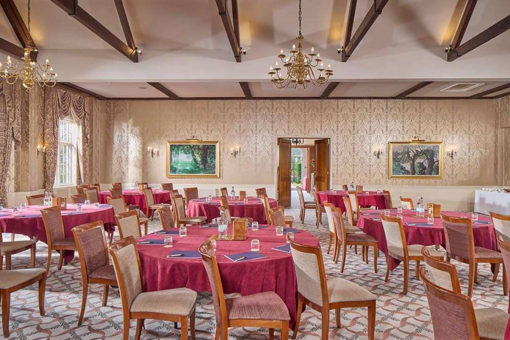 Elegant ballroom set with round tables dressed in burgundy linens, wooden chairs, crystal chandeliers, and formal place settings.