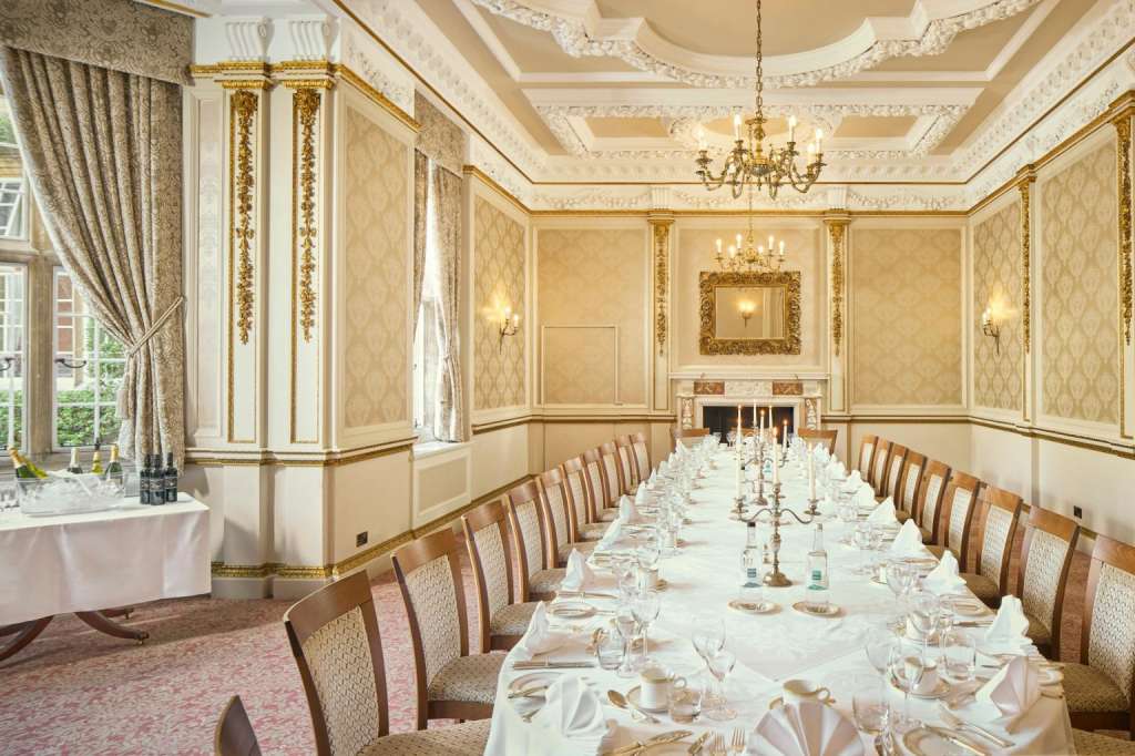 Formal dining room with long banquet table, place settings, wooden chairs, ornate ceiling, fireplace, and gold-trimmed wallpaper.