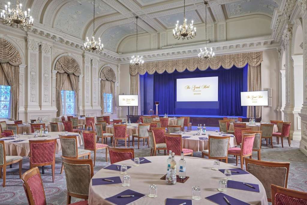 Elegant ballroom with round tables, red upholstered chairs, ornate ceiling, chandeliers, and blue stage with projection screen.