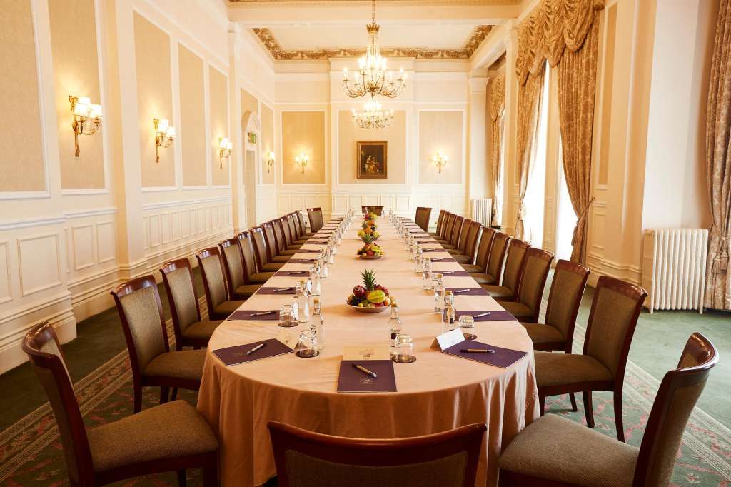 Elegant boardroom with long banquet table, formal place settings, gold drapes, and crystal chandeliers.
