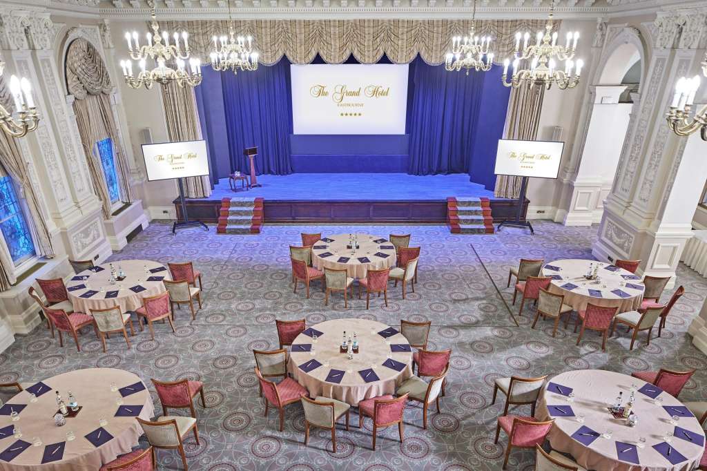 Grand ballroom with stage, blue curtains, crystal chandeliers, round tables with red chairs, and branded signage.