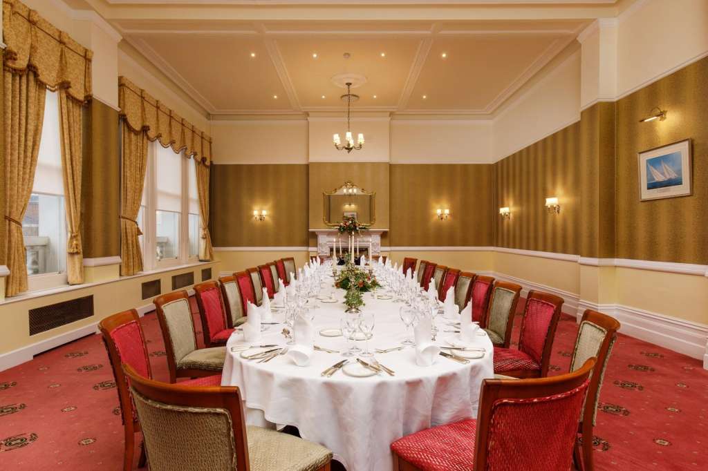 Formal dining room with long rectangular table set for seated dinner, red and cream chairs, gold drapes, and recessed ceiling lighting.