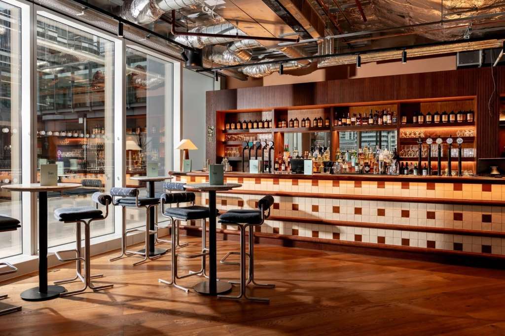 Modern bar interior with illuminated shelving, high-top seating, and floor-to-ceiling windows overlooking city.