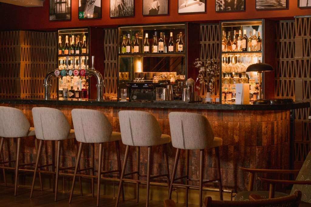 Bar counter with upholstered stools, illuminated bottle displays, and framed photographs on burgundy wall.