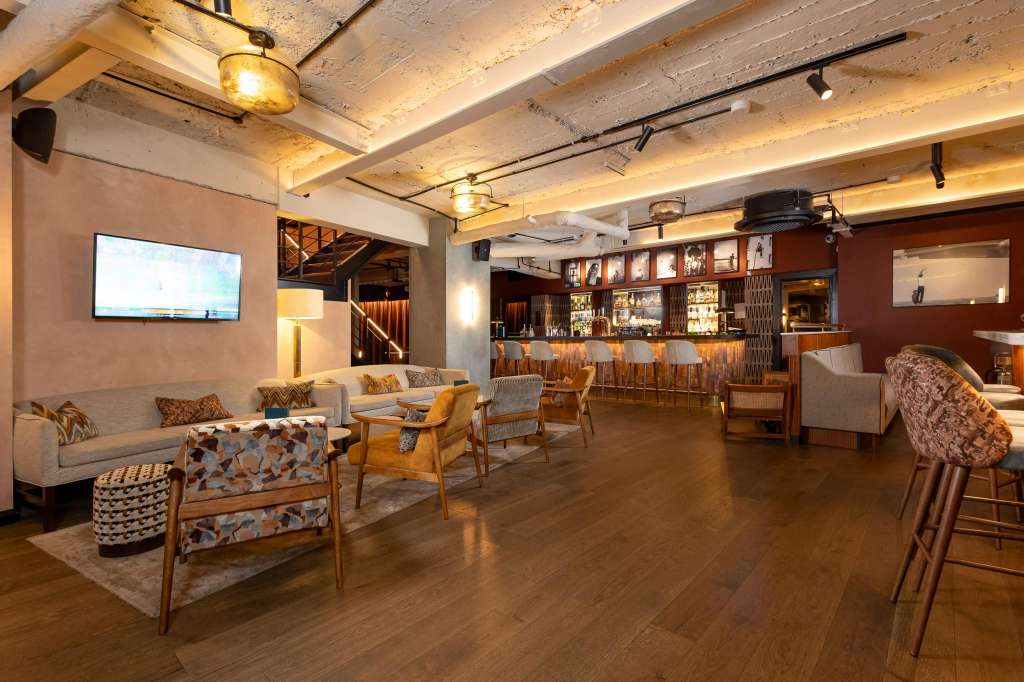 Modern lounge with exposed ceiling, warm lighting, seating areas, bar counter, and wall-mounted screen.