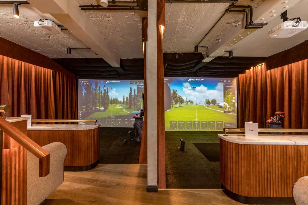 Indoor golf simulator space with two screens displaying golf courses, wooden curved counters, and orange curtains.