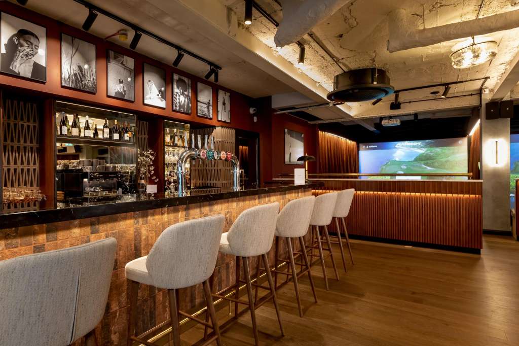 Bar area with upholstered seating, copper-tiled counter, stocked shelving, and digital display screen.