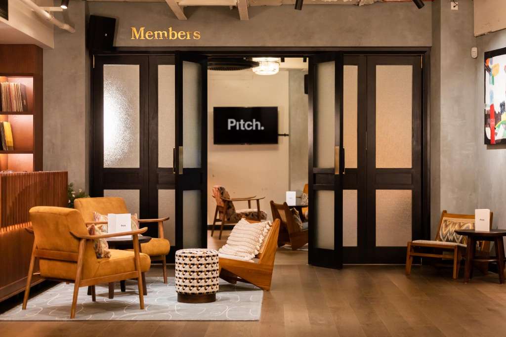 Members lounge with mid-century modern seating, wooden armchairs, patterned ottoman, and glass-fronted meeting room visible beyond.