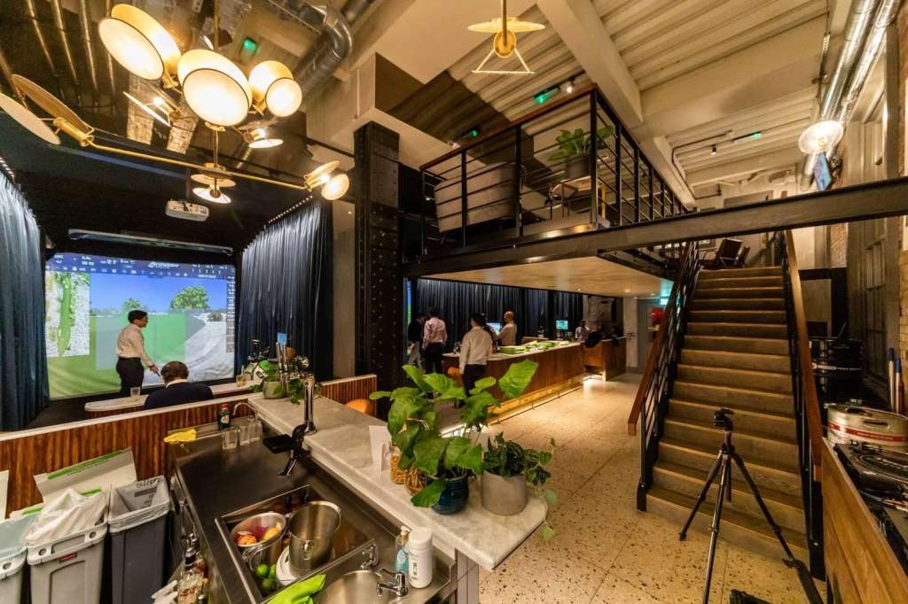 Modern event space with golf simulator screen, bar counter, mezzanine level, and warm lighting. Several people visible.