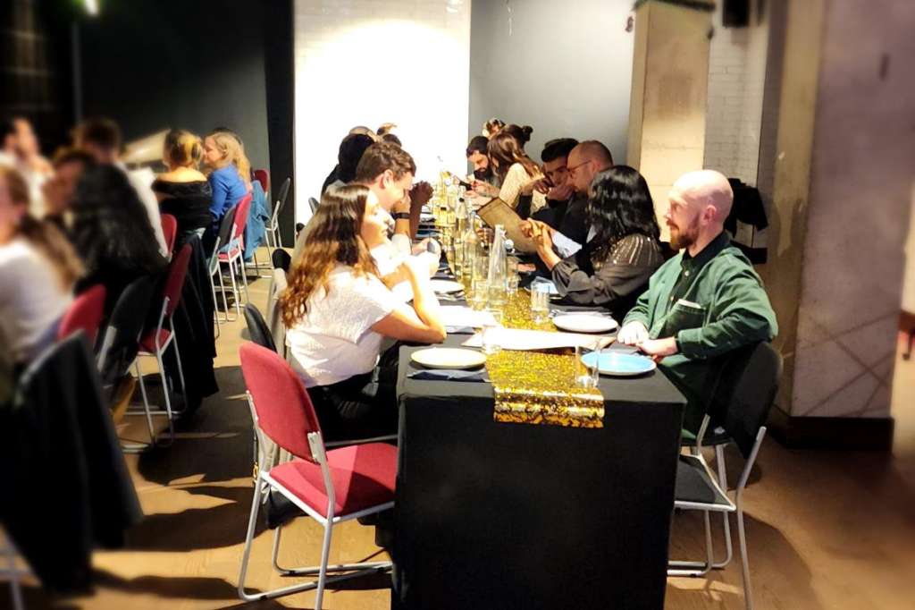 Indoor event space with guests seated at long tables with gold runners, modern lighting, and industrial ceiling.
