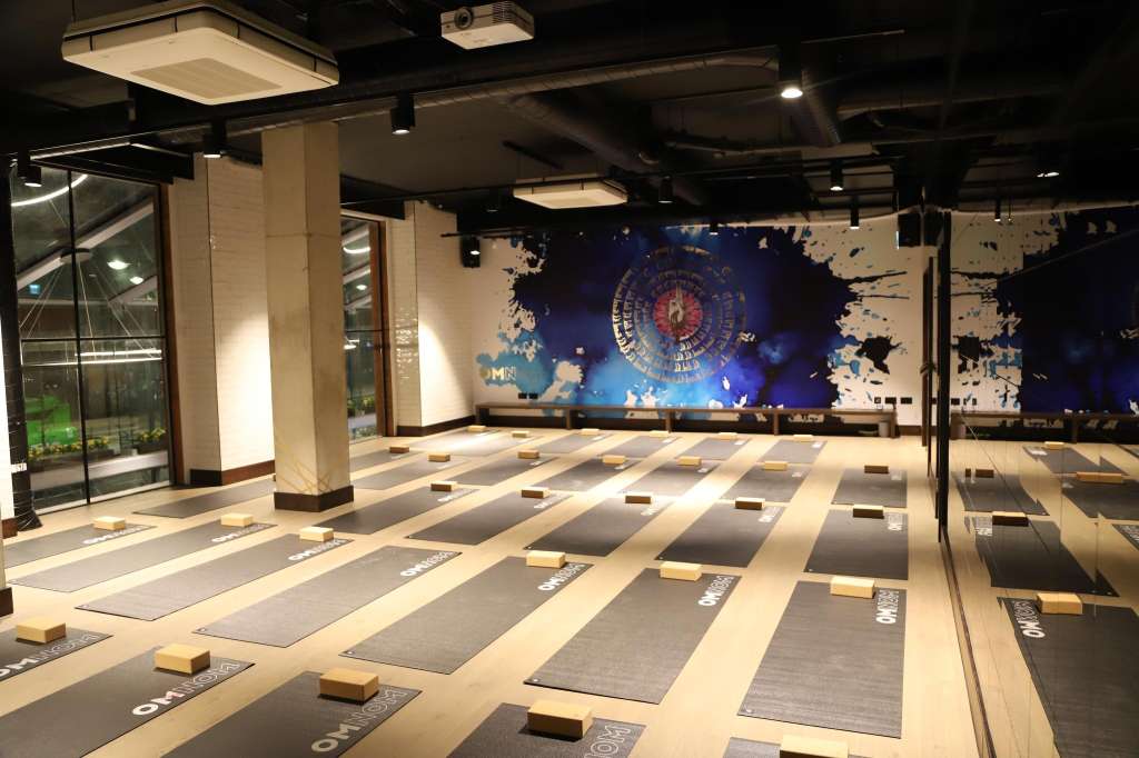Large open studio space with yoga mats arranged in rows, wooden blocks, and branded floor markers under modern ceiling lights.