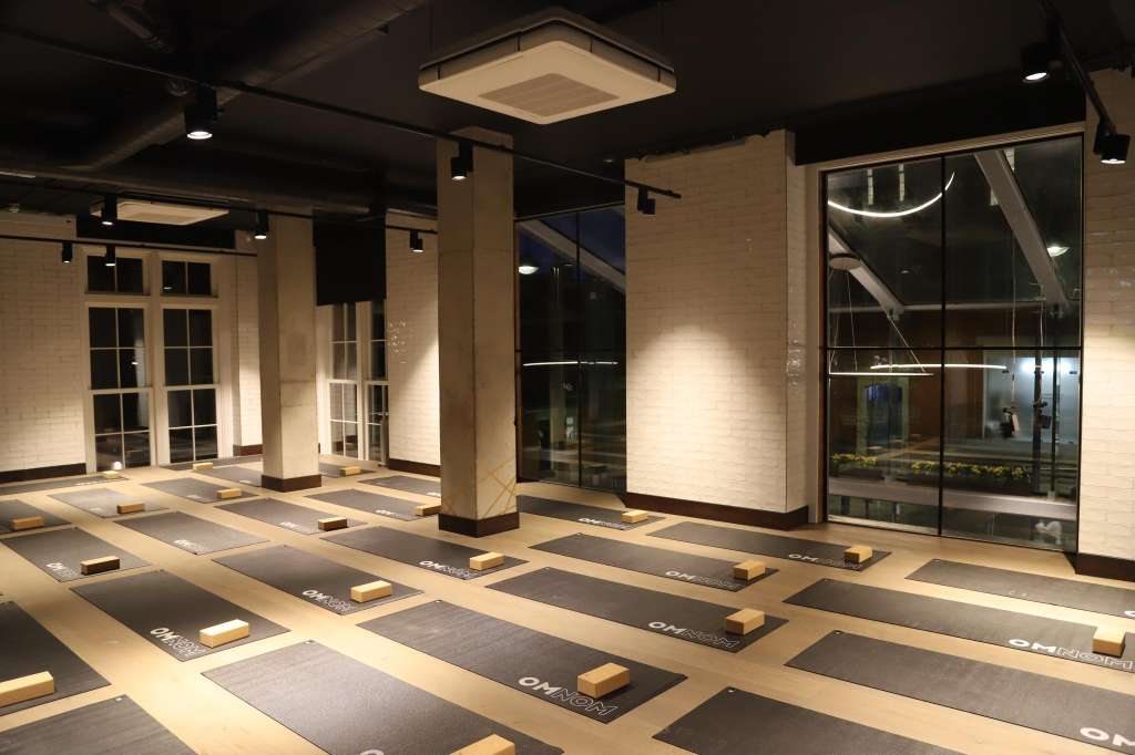 Modern studio space with yoga mats arranged in rows, wooden blocks, brick walls, and floor-to-ceiling windows.