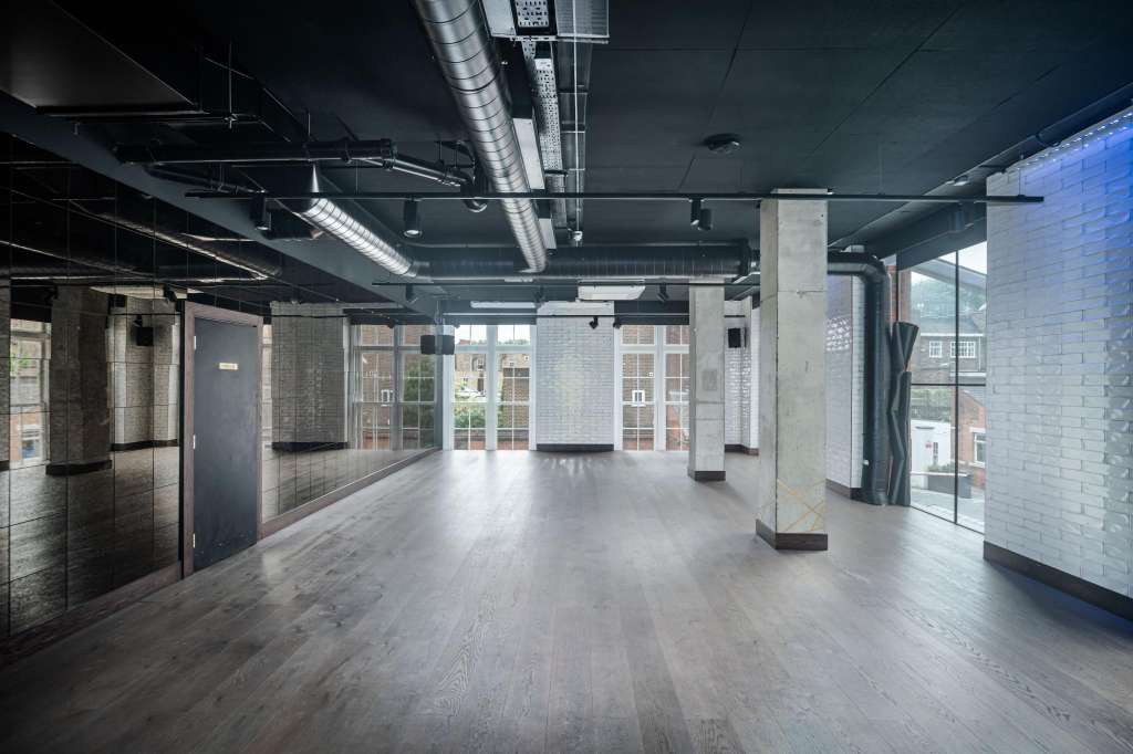 Industrial loft space with exposed ductwork, concrete columns, wooden flooring, and large windows overlooking urban buildings.