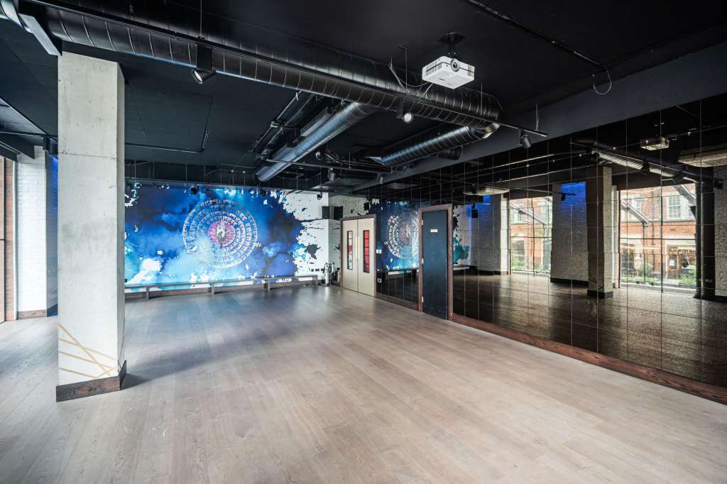 Modern industrial space with exposed ductwork, wooden flooring, and large blue digital display wall projection.