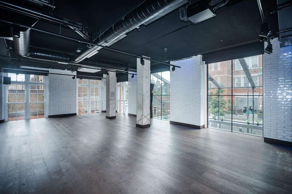 Empty industrial loft space with exposed ductwork, white brick walls, concrete columns, and large street-facing windows.