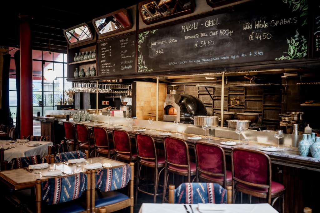 Restaurant dining area with counter seating, open kitchen, chalkboard menu, and set tables with patterned chair covers.