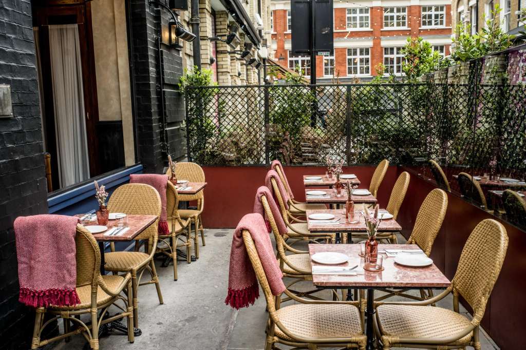 Outdoor terrace with bistro-style seating, set tables with place settings, pink accents, and climbing ivy on perimeter fencing.