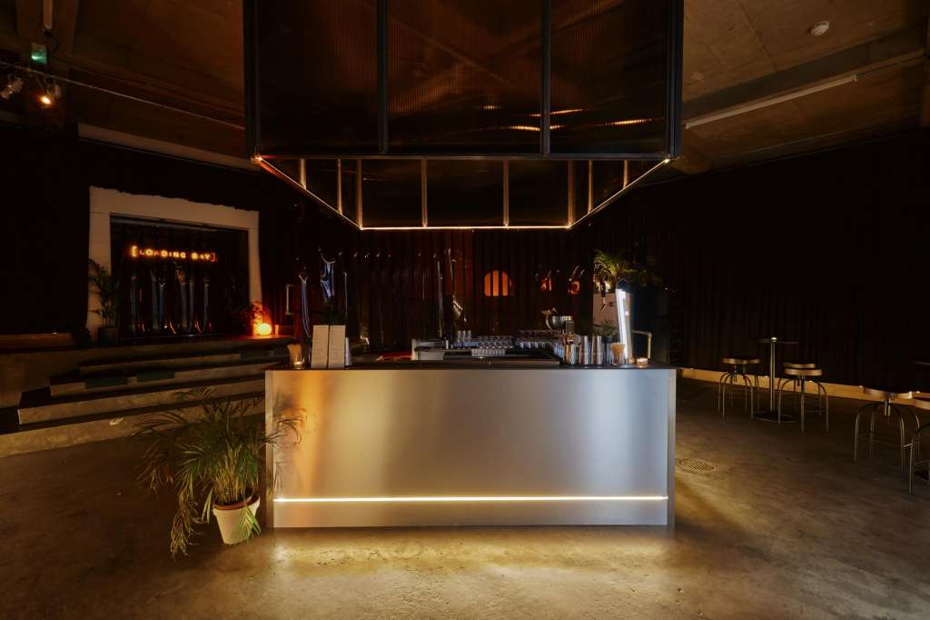 Modern bar setup with illuminated counter, neon signage, geometric ceiling lights, and potted plants in an industrial space.