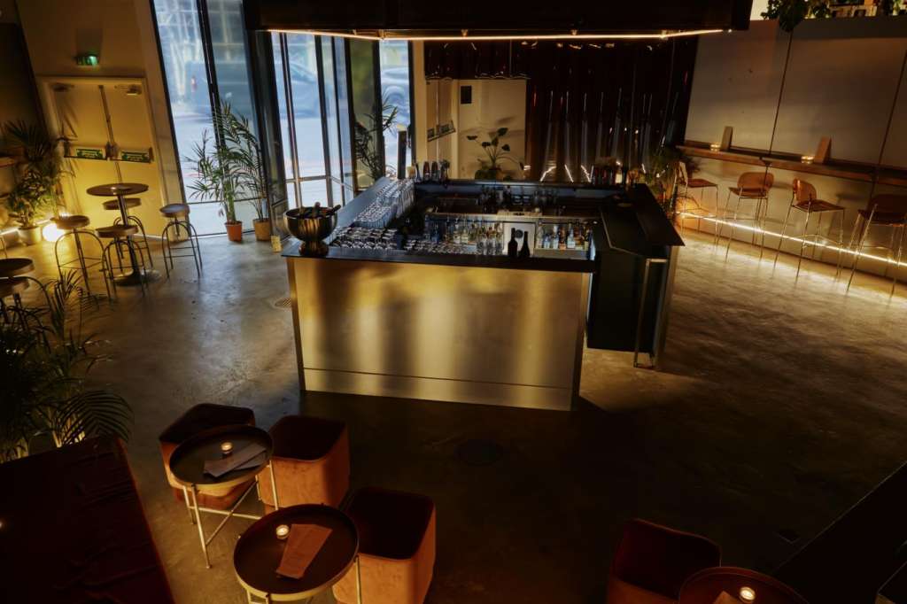 Industrial space with bar counter, high ceilings, pendant lighting, and scattered high-top tables with red seating.